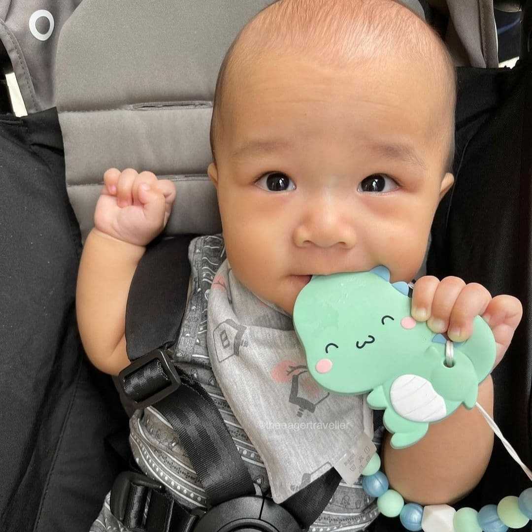 A baby sitting in a car seat holding a mint green dinosaur-shaped teether with a beaded strap.