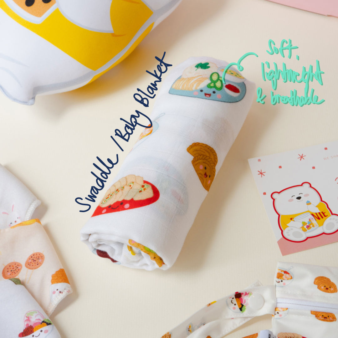 A soft baby swaddle blanket with a cute cartoon bear and food print, shown rolled up on a light cream background with other matching baby textiles nearby.