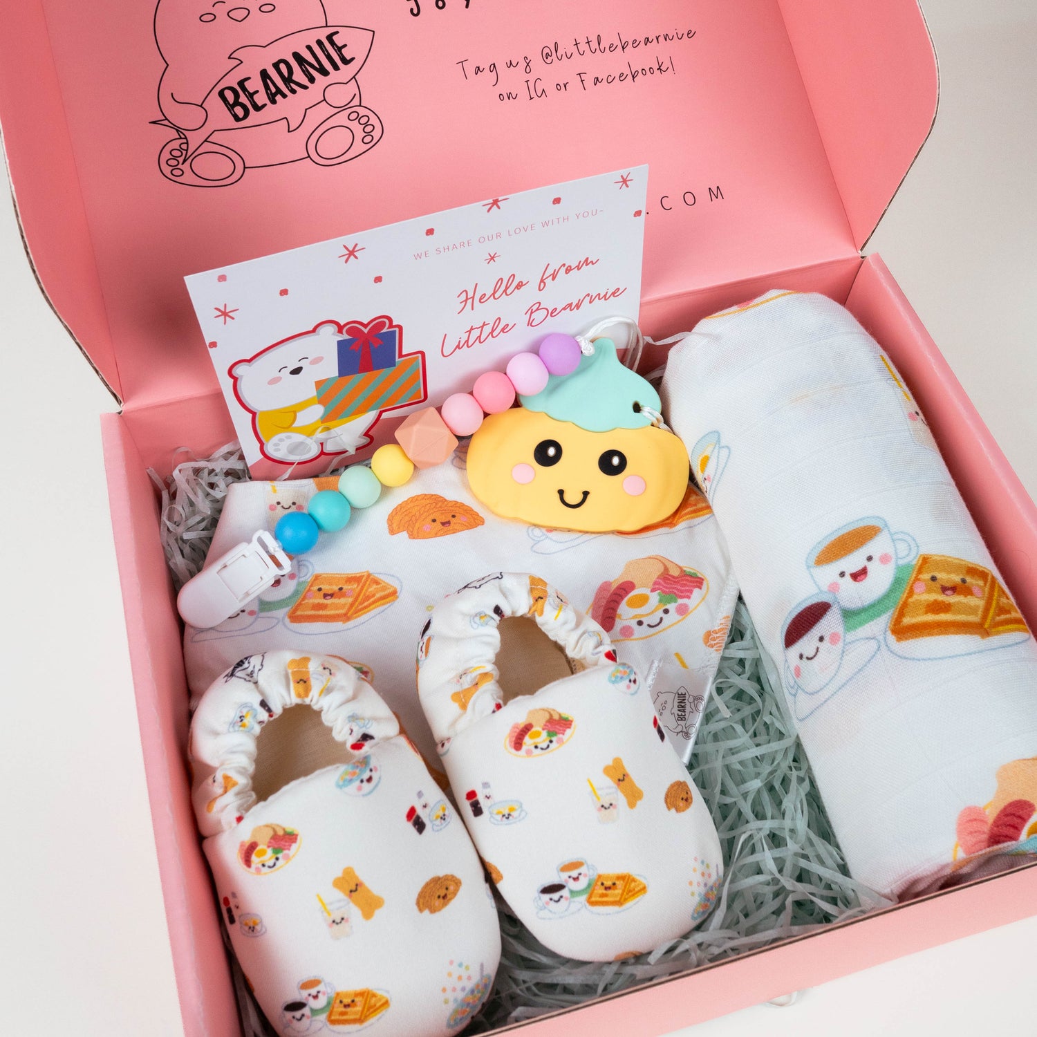 A pink gift box opened to reveal a baby gift set with matching printed items, including a swaddle wrap, booties, a teething toy, and a pacifier clip, arranged in shredded paper inside a branded Bearnie box.