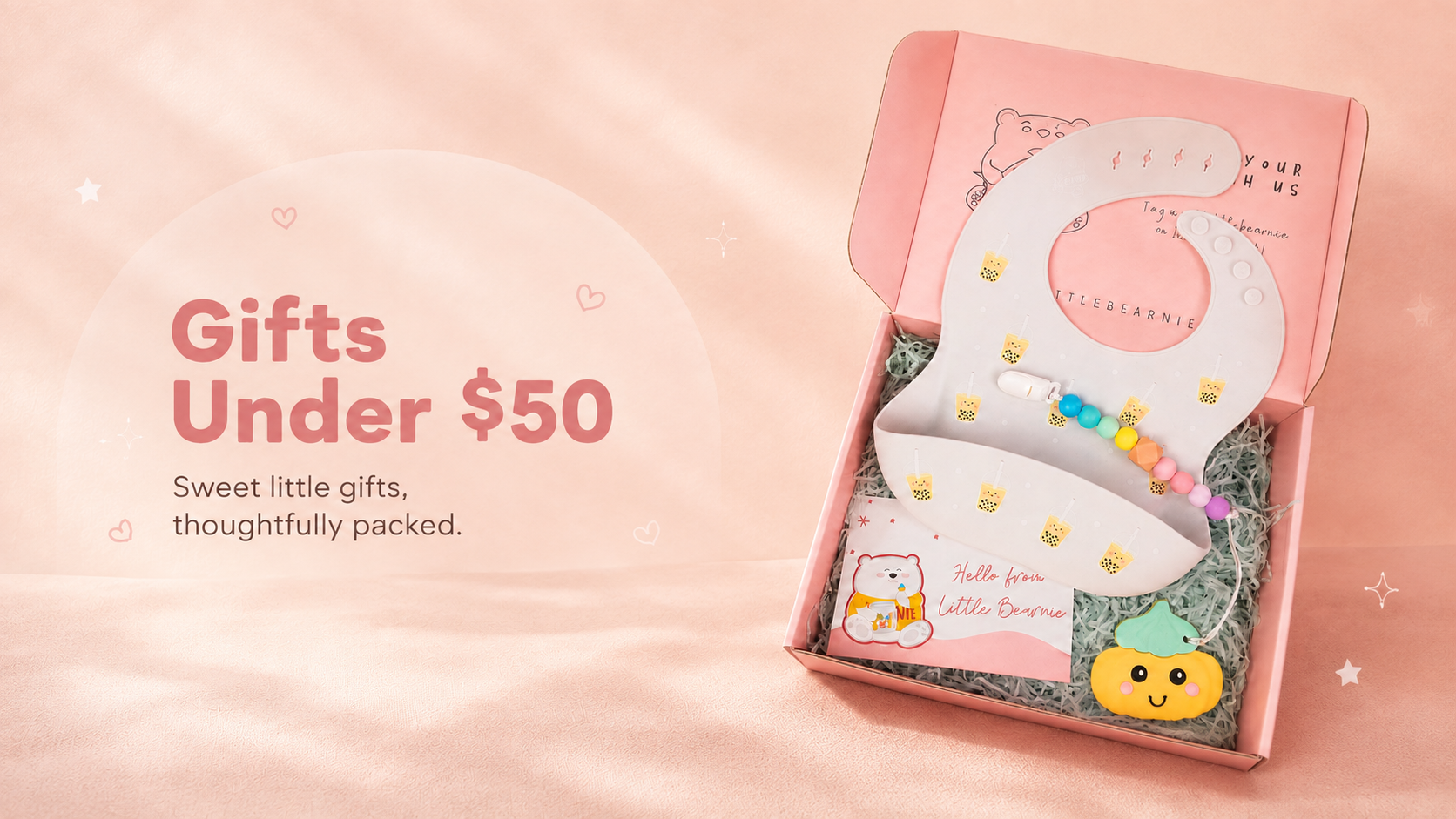 A pink gift box on a soft pink background containing a white baby bib with a pastel beaded bracelet and small gift items nestled in gray filler. Text on the left reads 'Gifts Under $50' with decorative hearts and stars.